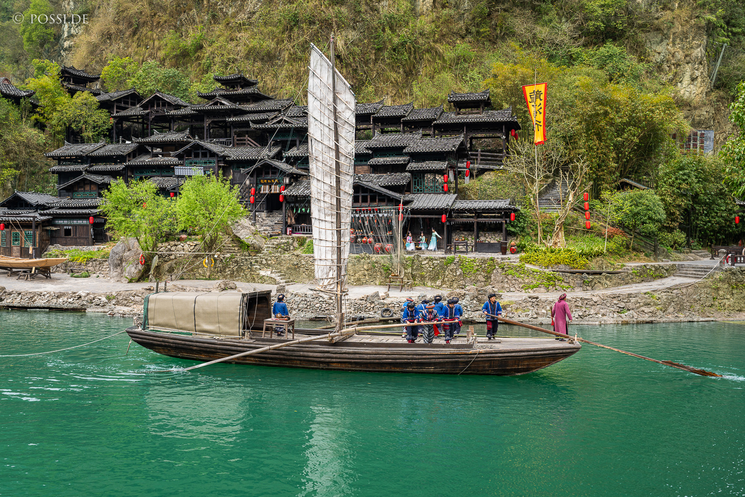 Das Bild zeigt ein traditionelles chinesisches Dorf am Ufer eines Flusses, umgeben von dichtem Grün und mit einem traditionellen Boot auf dem Wasser. (31.03.2025)
