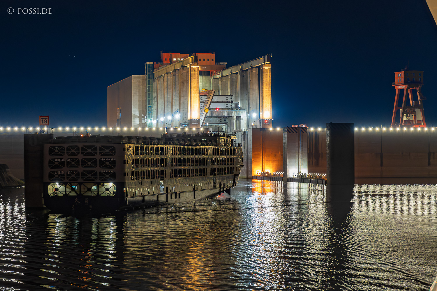 Das Bild zeigt eine Industrieanlage am Wasser, die bei Nacht beleuchtet ist und einen industriellen Charakter ausstrahlt. (31.03.2025)
