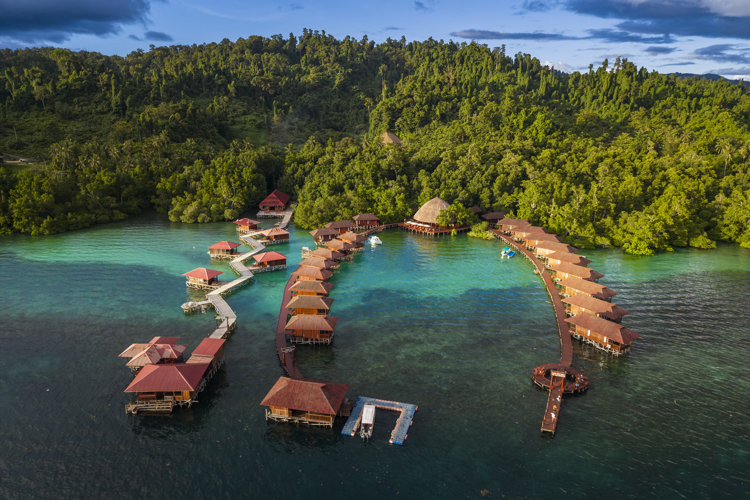 Luftaufnahme hölzerner Wasserbungalows, die in das türkisfarbene Wasser von Raja Ampat hineinragen, vor dichtem tropischem Regenwald. West Papua 2025. (04.10.2025)