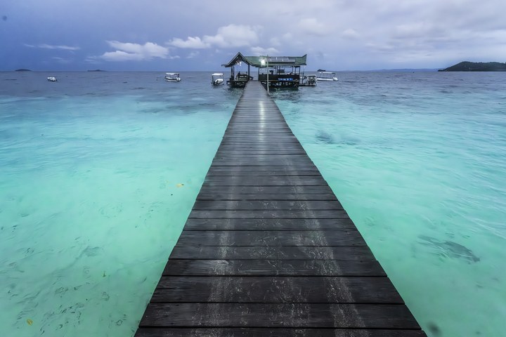 Ein langer Holzsteg führt über türkisfarbenes Wasser zu einem Bau mit traditionellem Dach. West Papua 2025. (09.10.2025)